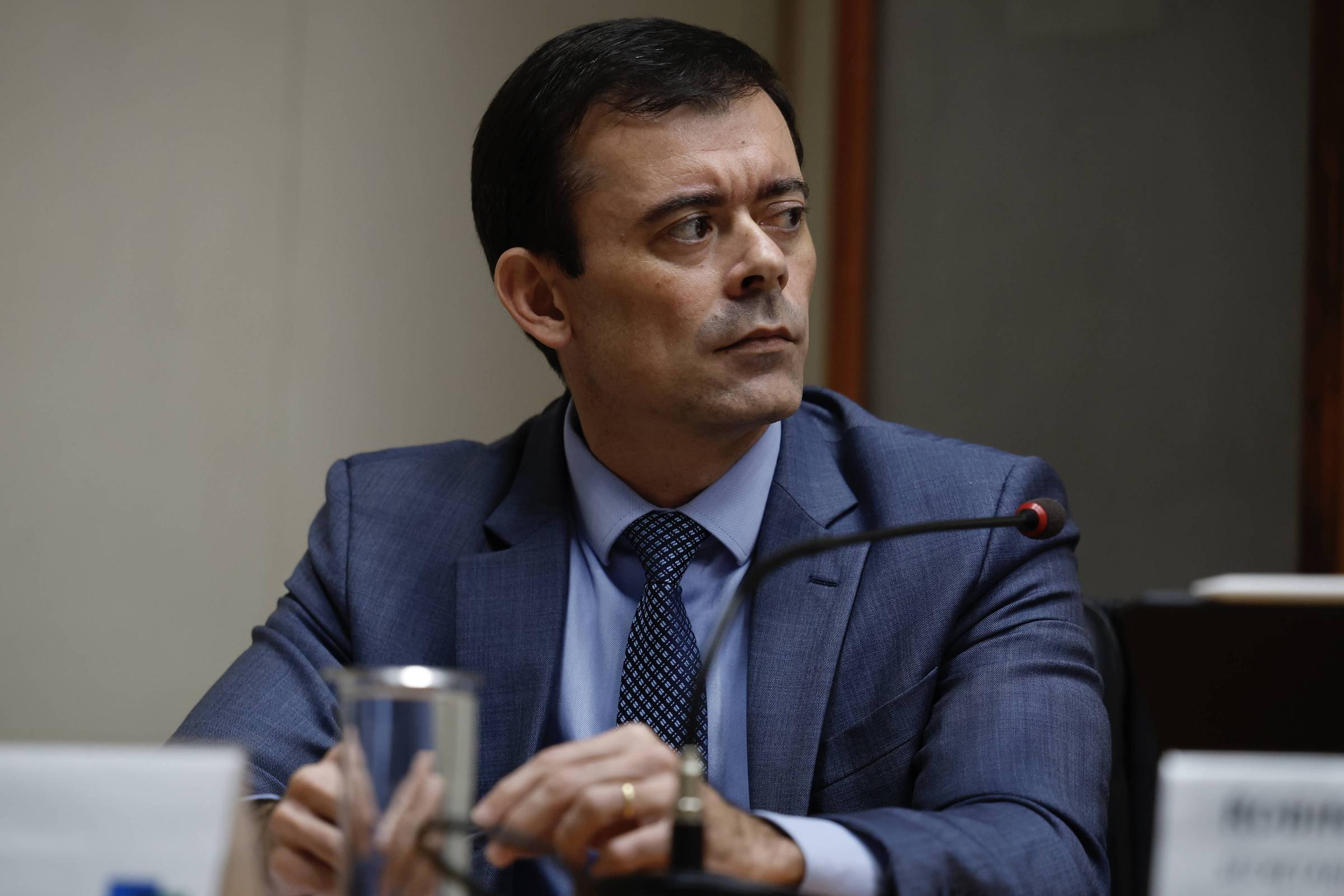 Um homem está sentado em uma mesa, vestindo um terno azul. Ele tem cabelo curto e está olhando para o lado com uma expressão séria. Na mesa, há um copo. O fundo é neutro, com uma parede clara.