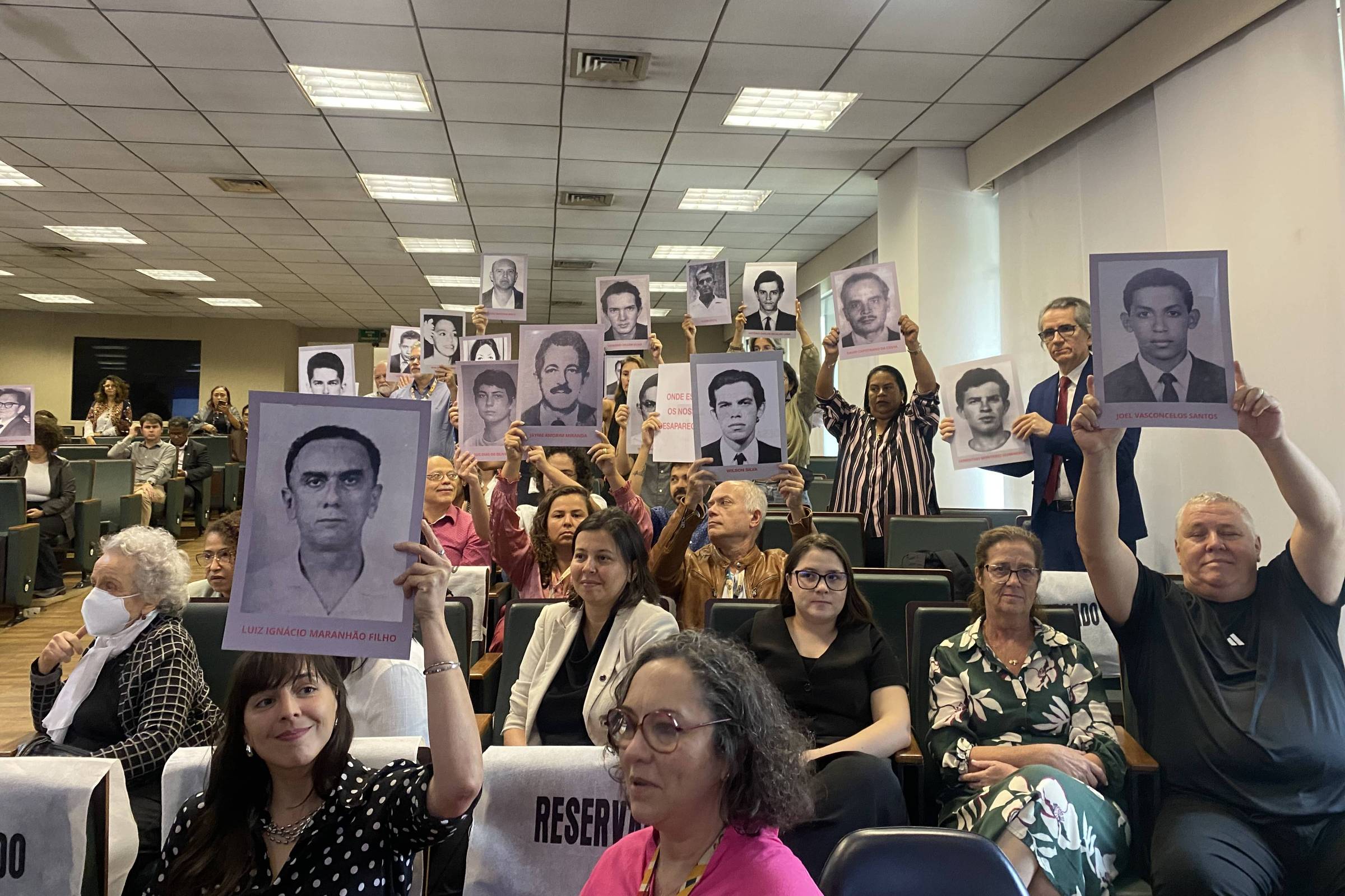A imagem mostra um grupo de pessoas em um auditório, segurando cartazes com retratos de homens. As pessoas estão sentadas e algumas estão em pé, levantando os cartazes. O ambiente é bem iluminado, com mesas e cadeiras organizadas. Há uma variedade de expressões faciais entre os participantes, que parecem estar em um evento significativo.