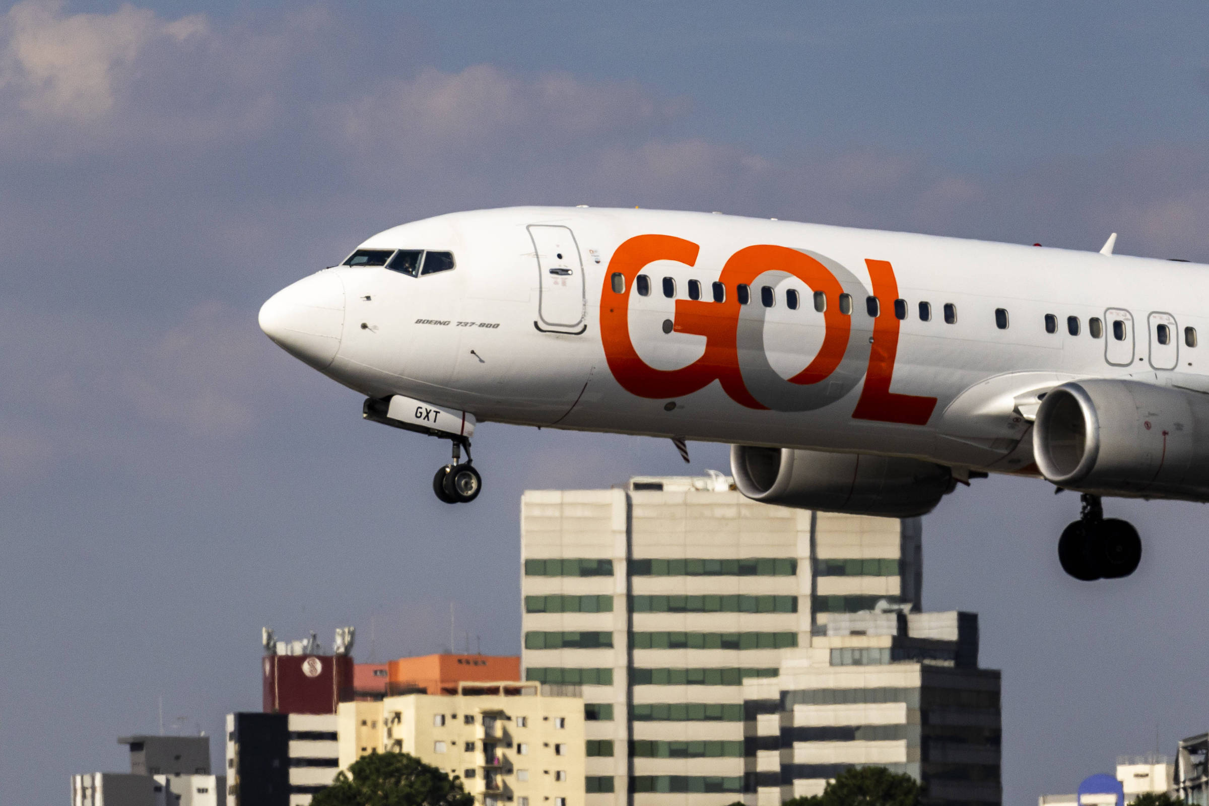Um avião da companhia aérea GOL, modelo Boeing 737, está em fase de pouso, com a fuselagem branca e o logotipo laranja da empresa bem visível. Ao fundo, há uma cidade com vários edifícios, alguns deles de diferentes alturas e cores, sob um céu parcialmente nublado.