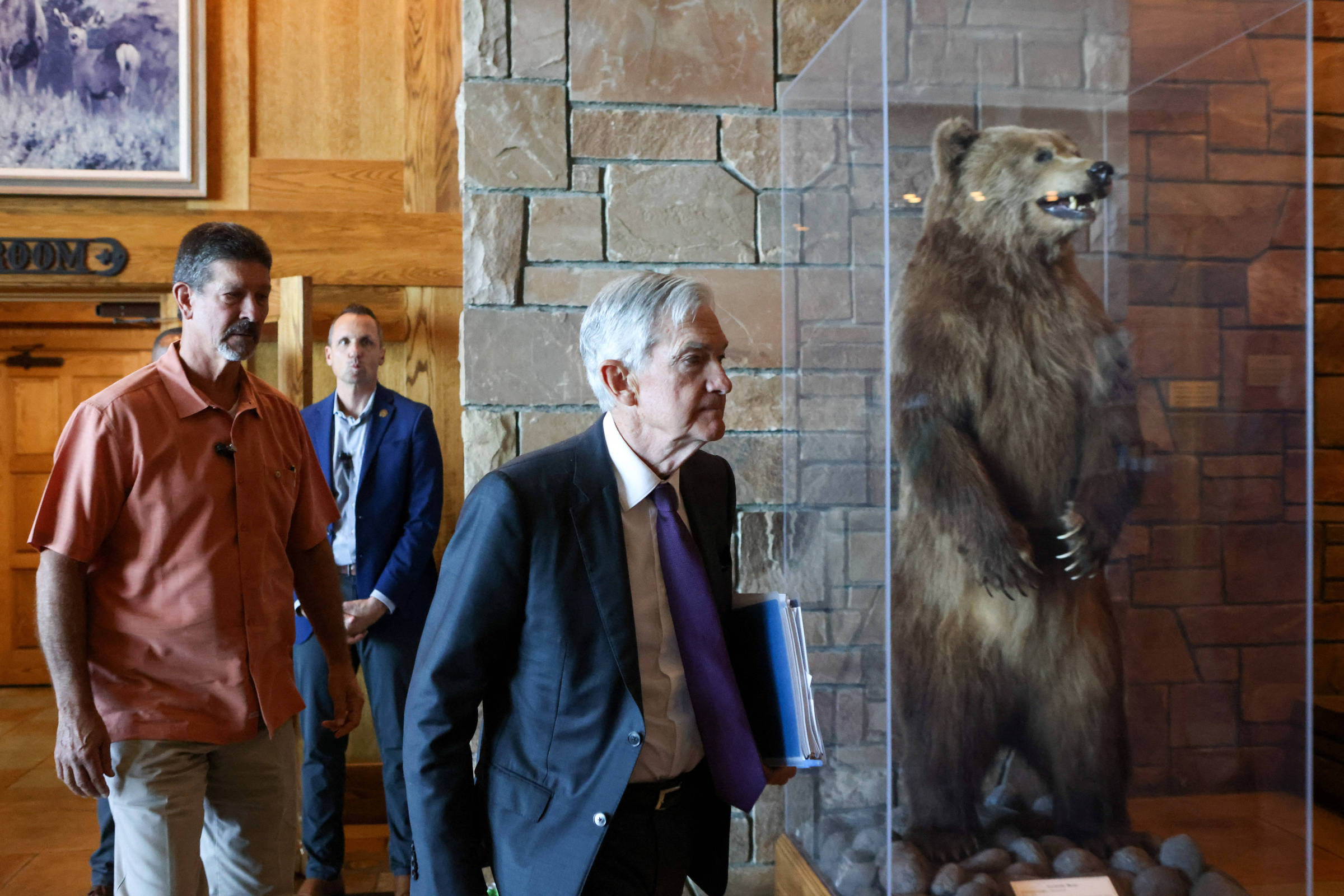 A imagem mostra três homens em um ambiente interno, possivelmente um museu ou uma sala de exposições. Um dos homens, com cabelo grisalho e terno escuro, está caminhando em direção a uma vitrine que contém um urso empalhado. Ao fundo, há um homem com uma camisa laranja e outro com um terno azul. As paredes são de pedra e há uma decoração rústica ao redor.