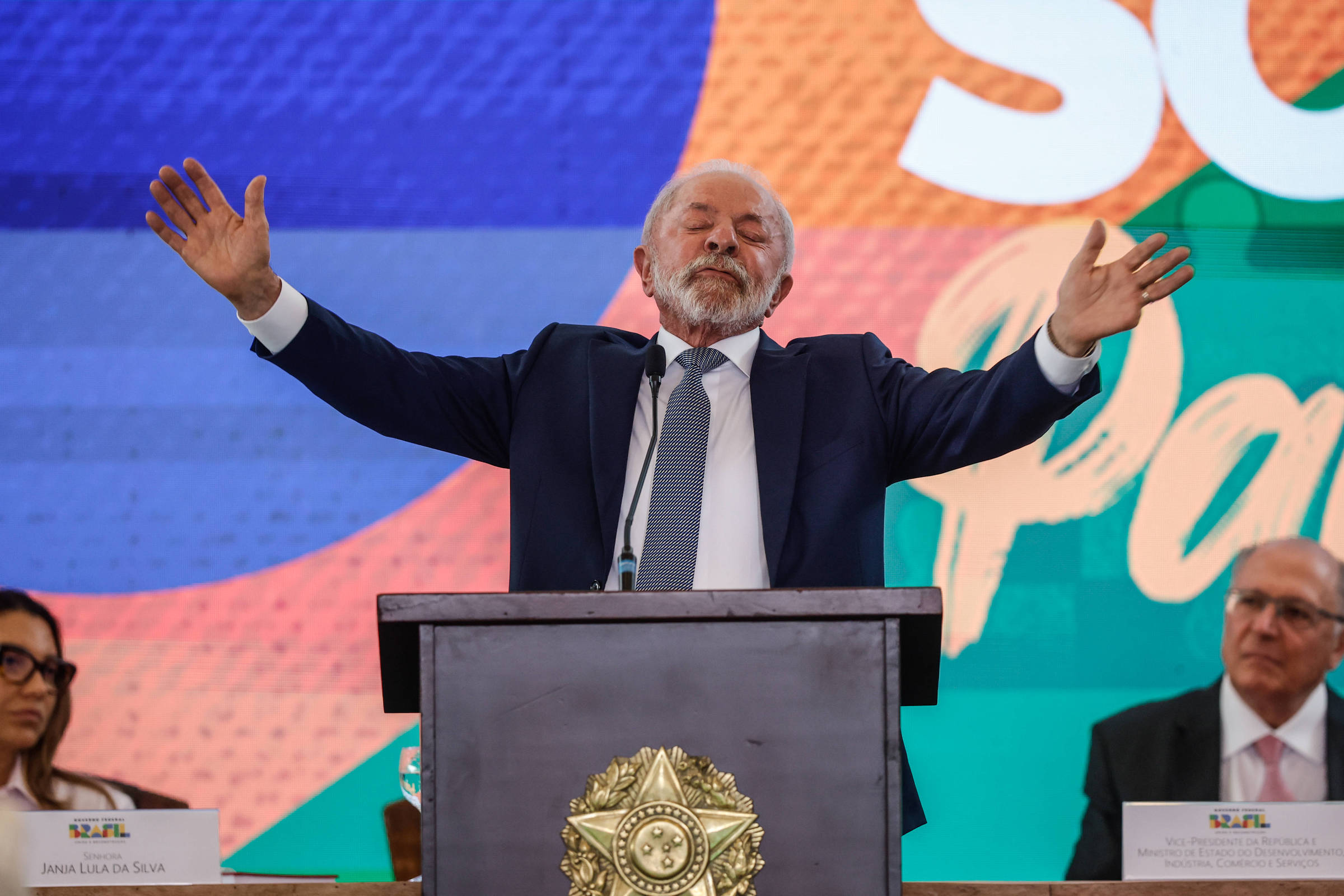 Um homem de terno e gravata, com cabelo grisalho e barba, está em pé atrás de um púlpito, gesticulando com os braços abertos. Ele parece estar fazendo um discurso em um evento oficial. Ao fundo, há um painel colorido com as palavras