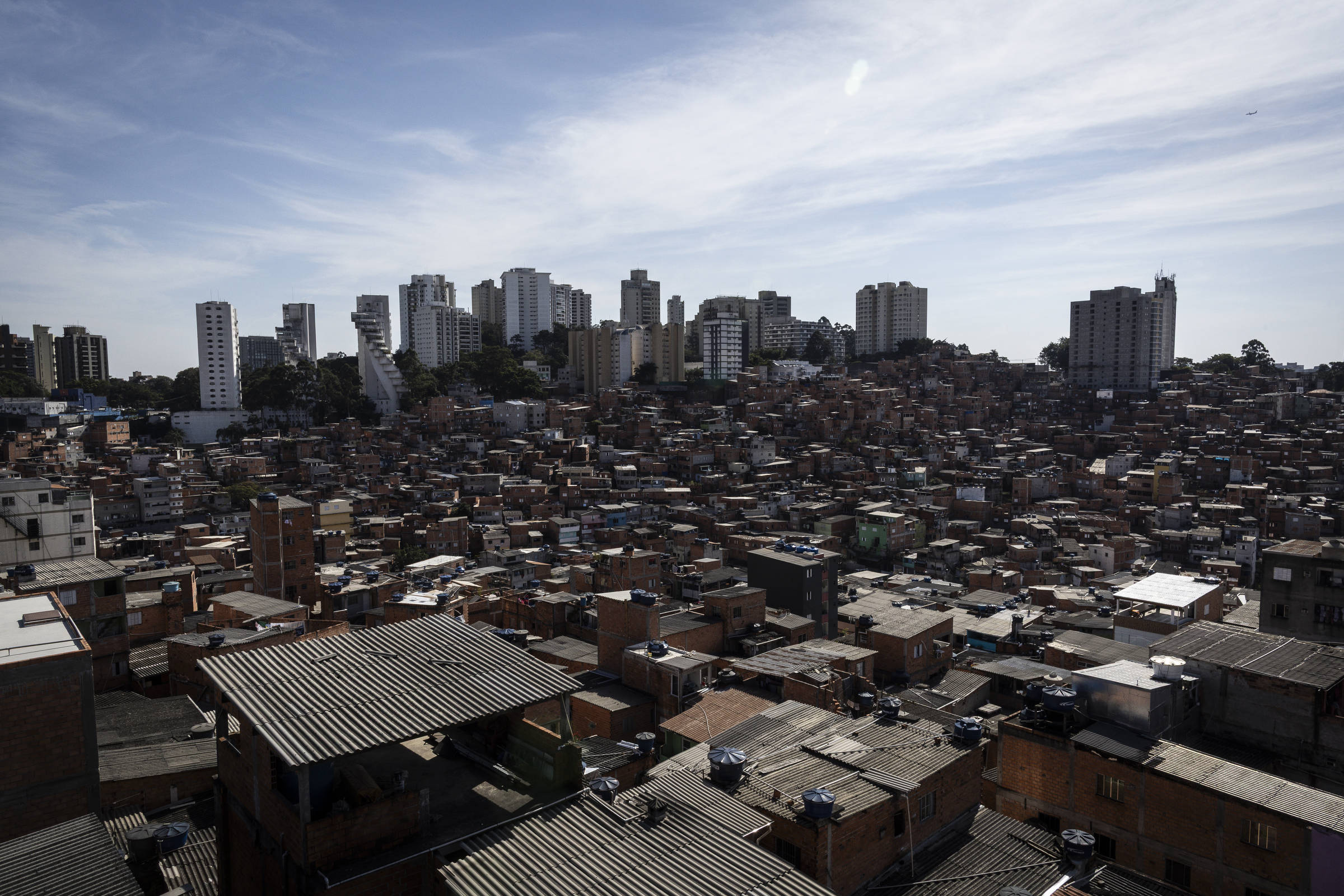 A imagem mostra uma vista panorâmica de uma comunidade urbana, com uma densa concentração de casas de tijolos e telhados de metal. Ao fundo, há prédios altos e modernos, contrastando com a arquitetura simples das casas. O céu está parcialmente nublado, e a cena reflete uma mistura de áreas de habitação precária e desenvolvimento urbano.