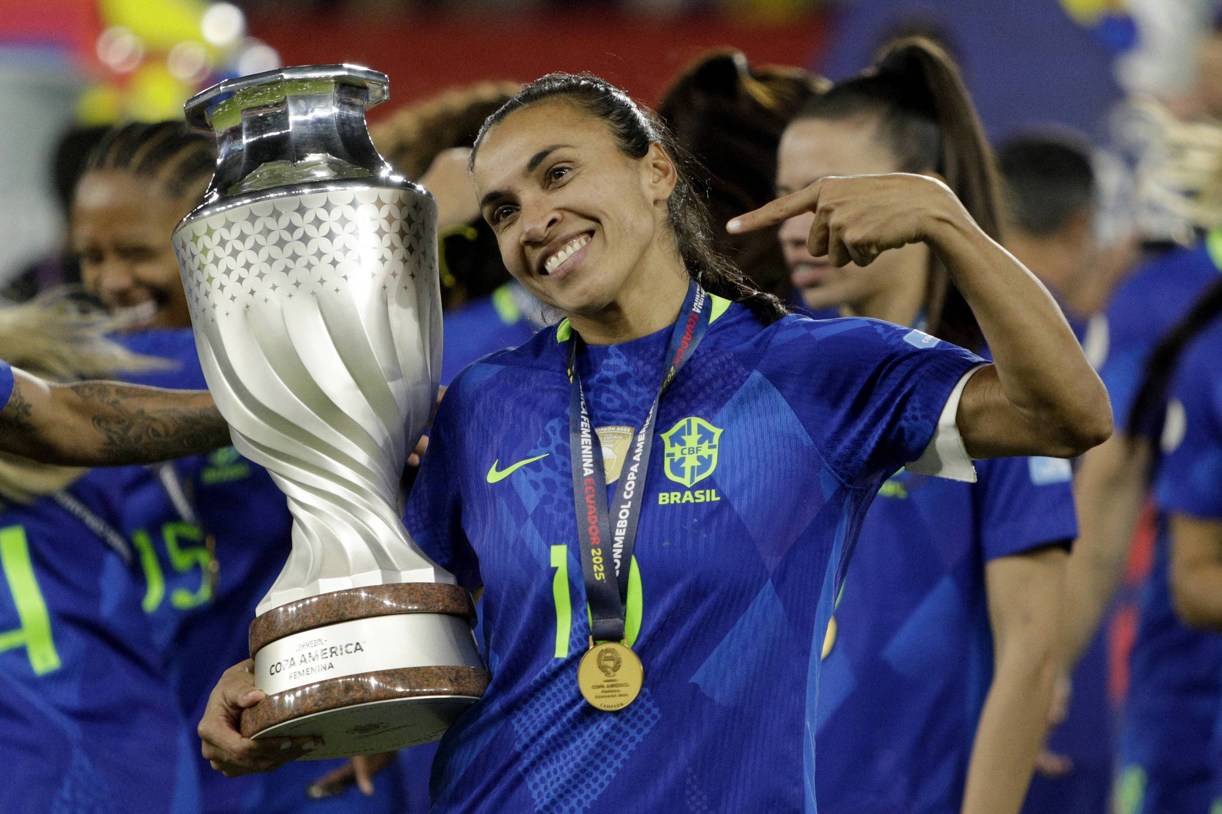 Uma jogadora de futebol vestindo um uniforme azul do Brasil, segurando um troféu grande e brilhante. Marta está sorrindo e fazendo um gesto com a mão, apontando para si mesma. Ao fundo, outras jogadoras também estão celebrando. A cena parece ser de uma cerimônia de premiação.
