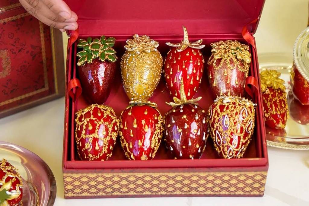 Uma caixa vermelha aberta contendo morangos decorativos. Os morangos são de cores vibrantes, com detalhes dourados e verdes, e estão dispostos em um forro vermelho. Um dos morangos tem uma cobertura dourada, enquanto outros têm detalhes variados. Ao lado da caixa, há um prato com mais morangos decorativos.