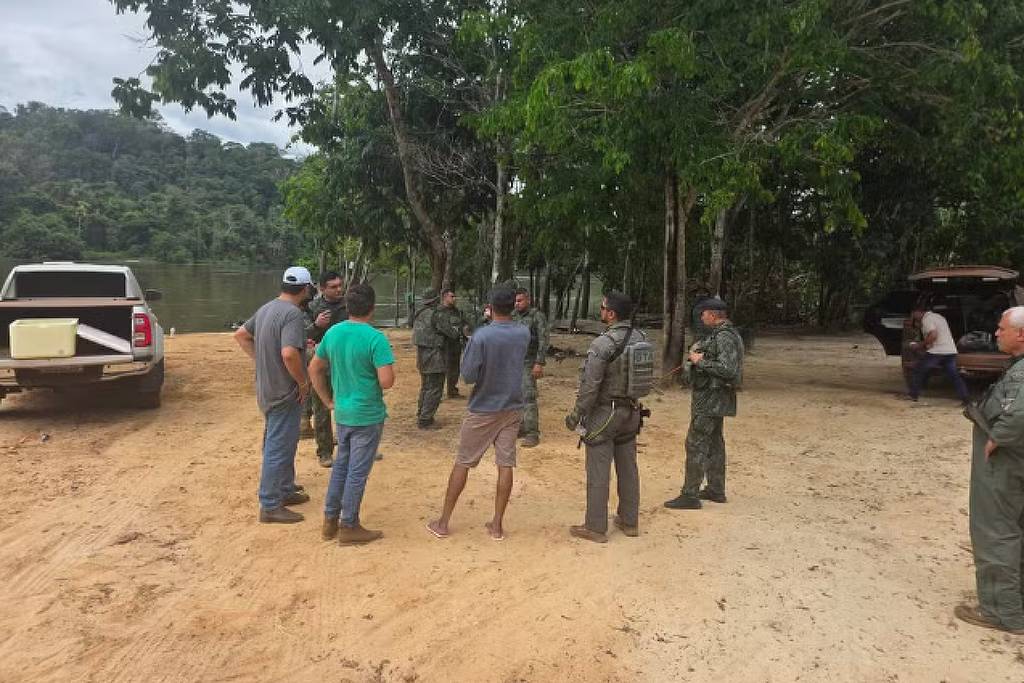 Um grupo de pessoas se reúne em uma área de areia próxima a um rio, cercada por árvores. Algumas pessoas estão vestidas com uniformes militares, enquanto outras estão em roupas casuais. Há veículos estacionados ao fundo, incluindo uma caminhonete. O ambiente é natural, com vegetação densa ao redor.