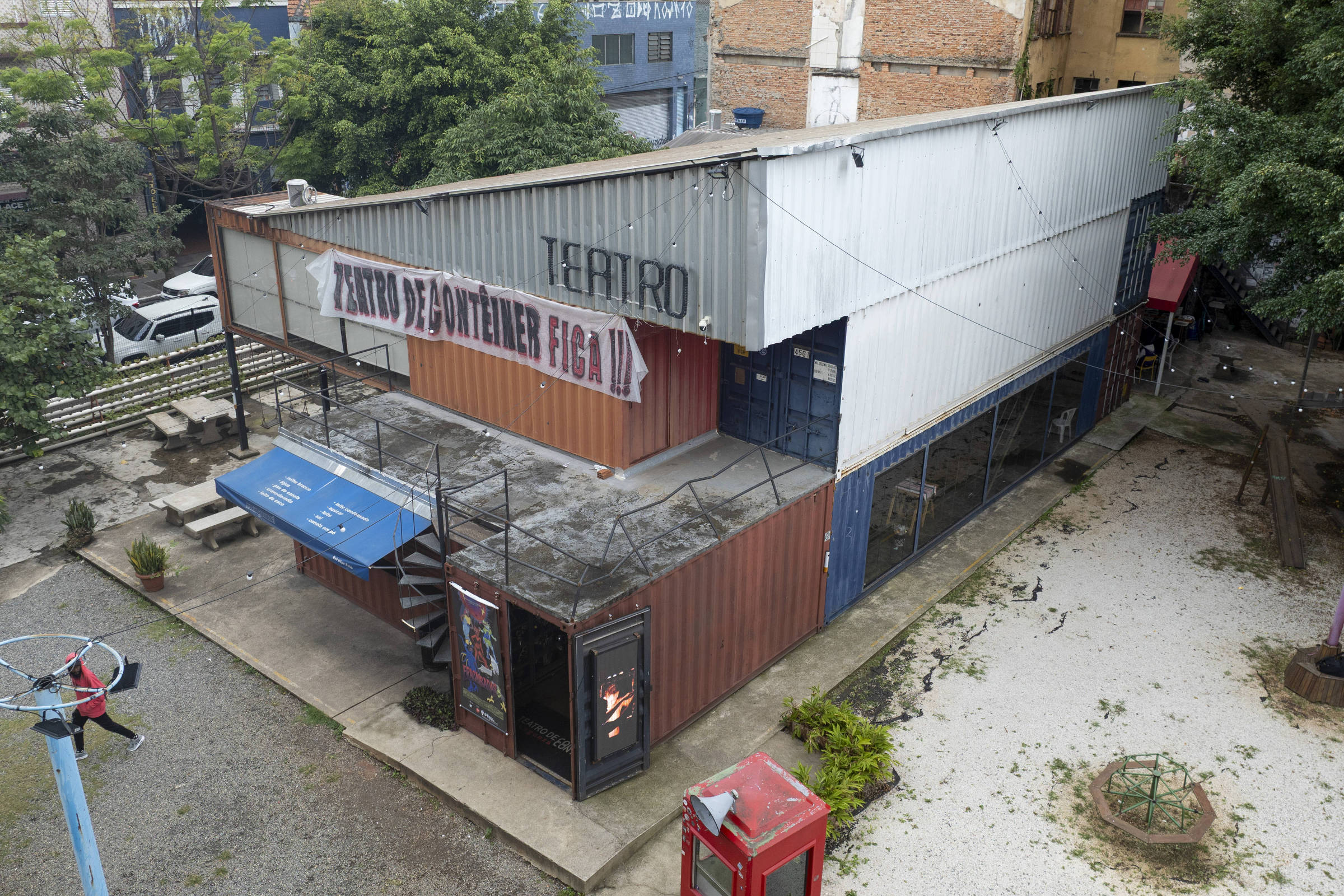 A imagem mostra um teatro localizado em uma área urbana. O edifício é de estrutura metálica e possui um telhado de duas águas. Na fachada, há uma placa com o nome do teatro, que é parcialmente visível. O local parece ter um espaço ao ar livre ao redor, com algumas árvores e um parque infantil próximo. O chão é coberto por pedras e há um telefone público vermelho na parte inferior direita da imagem.