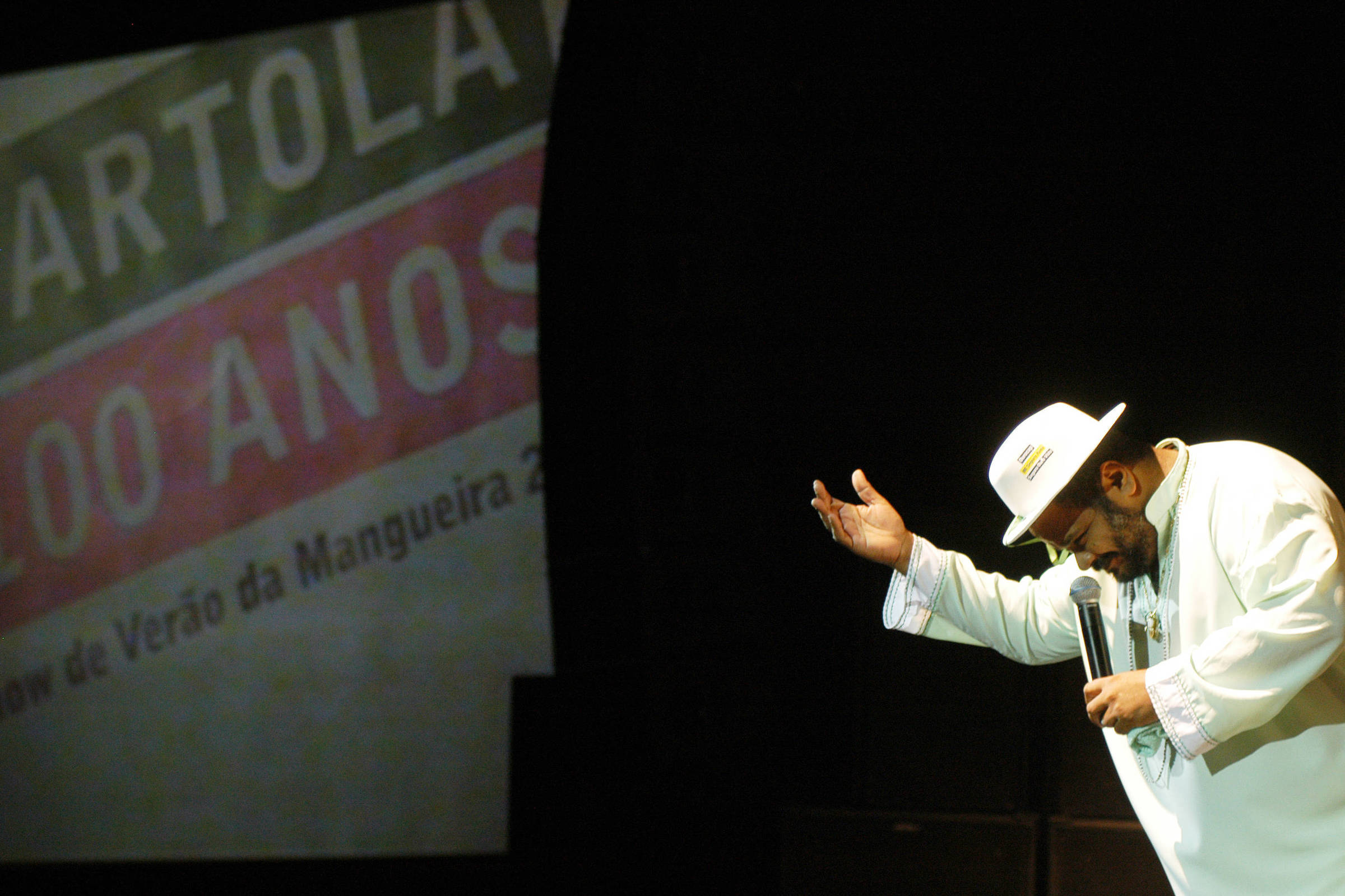 Um cantor se apresenta em um palco, usando um traje branco e um chapéu. Ele está gesticulando com a mão direita enquanto segura um microfone com a mão esquerda. Ao fundo, há um projetor exibindo a imagem com o texto