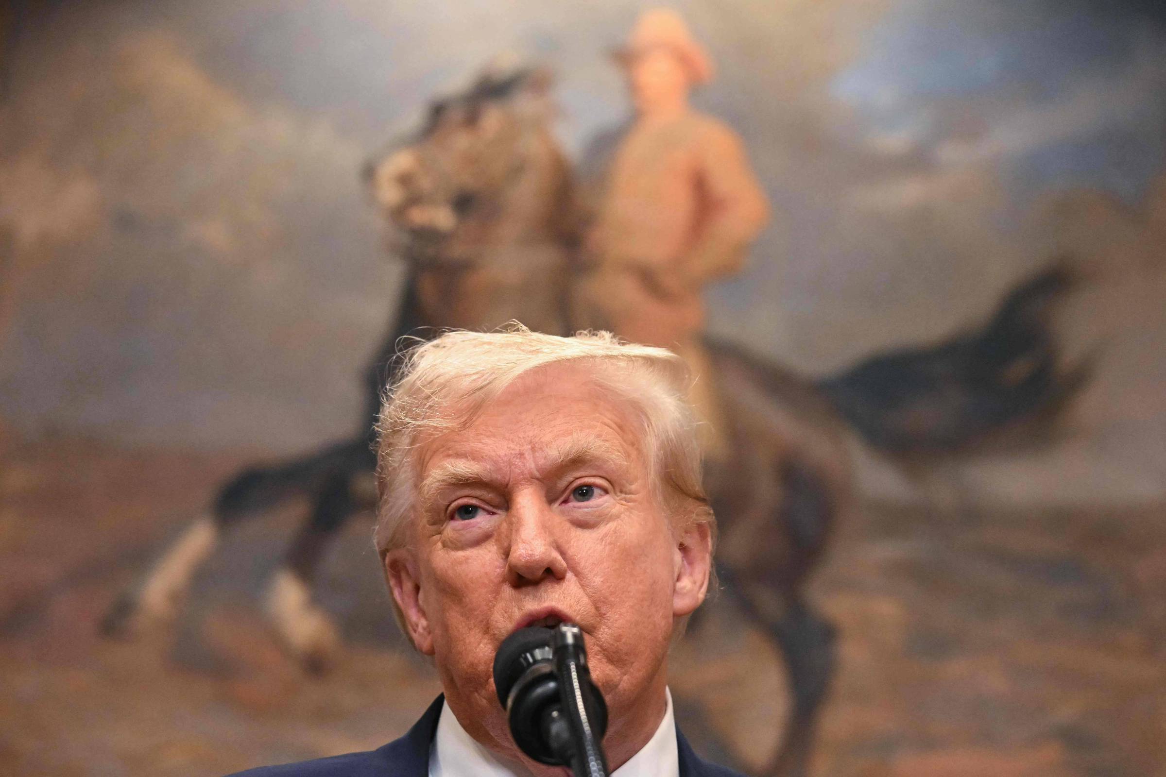 A imagem mostra um homem falando ao microfone, com cabelo loiro e pele clara. Ao fundo, há uma pintura de um homem montado em um cavalo, em um cenário que parece ser uma paisagem. O homem em primeiro plano está vestido com um terno escuro e uma camisa branca.