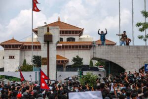 A imagem mostra uma grande multidão reunida em frente ao Parlamento do Nepal, com pessoas levantando as mãos e bandeiras do Nepal. No fundo, é possível ver a arquitetura do edifício do Parlamento, que possui um telhado característico. Algumas pessoas estão em uma estrutura elevada, enquanto outras estão na base, criando um ambiente de protesto e agitação. O céu está parcialmente nublado, e há árvores ao redor.