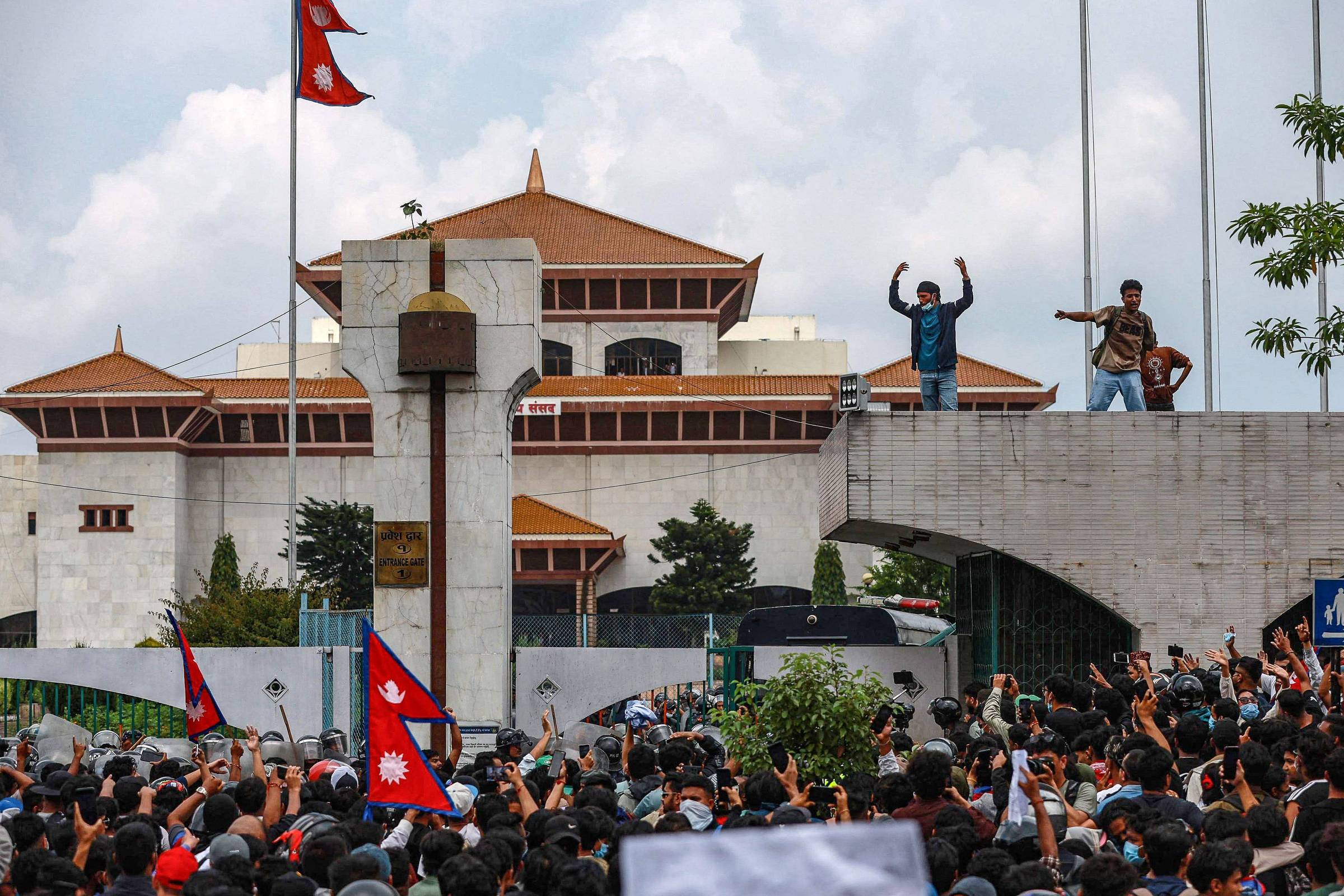 A imagem mostra uma grande multidão reunida em frente ao Parlamento do Nepal, com pessoas levantando as mãos e bandeiras do Nepal. No fundo, é possível ver a arquitetura do edifício do Parlamento, que possui um telhado característico. Algumas pessoas estão em uma estrutura elevada, enquanto outras estão na base, criando um ambiente de protesto e agitação. O céu está parcialmente nublado, e há árvores ao redor.