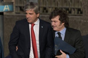 Na imagem, dois homens estão em pé, conversando. O homem à esquerda está usando um terno escuro com uma gravata vermelha e uma camisa branca, enquanto o homem à direita está vestido com um terno preto e uma camisa azul clara. Ambos parecem estar em um ambiente externo, com uma estrutura ao fundo que sugere um local urbano. O homem à direita segura um documento ou pasta em suas mãos.