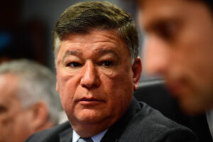 Um homem com cabelo curto e grisalho, usando um terno escuro e uma gravata, está sentado em uma mesa. Ele tem uma expressão séria e está olhando para o lado, enquanto outra pessoa está parcialmente visível ao seu lado. O fundo é desfocado, sugerindo um ambiente de reunião ou conferência.