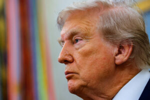 A imagem mostra um homem de perfil, com cabelo loiro e pele clara. Ele está com uma expressão séria, olhando para o lado. O fundo é desfocado, mas há cores que sugerem um ambiente interno, possivelmente um escritório ou sala de reuniões.