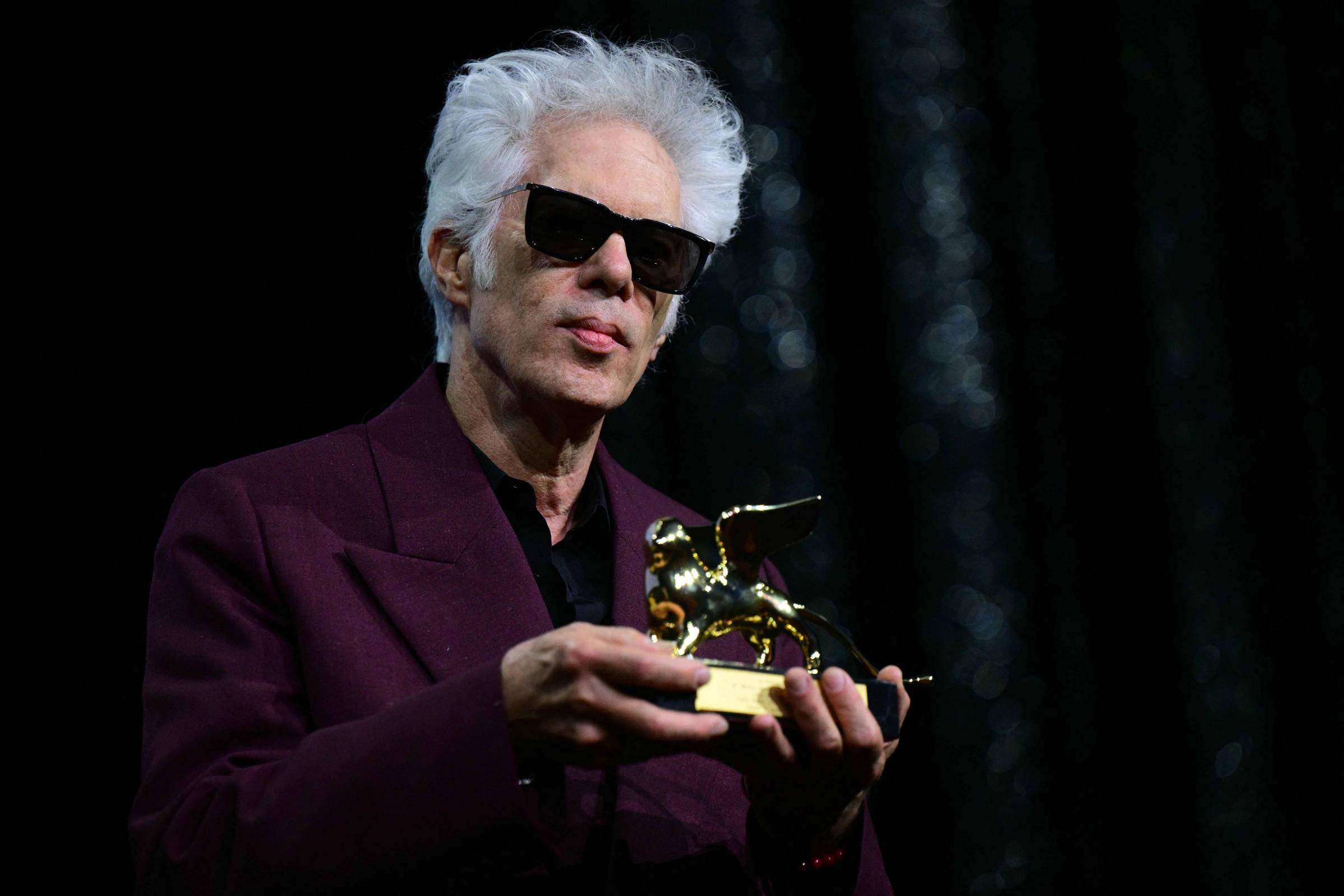 Um homem com cabelo grisalho e óculos escuros está segurando um prêmio dourado em forma de leão. Ele usa um paletó roxo e está em um palco com um fundo escuro. A expressão do homem é séria e ele parece estar recebendo o prêmio em um evento de premiação.