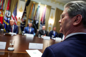 A imagem mostra uma reunião no Salão Oval da Casa Branca. Em primeiro plano, um homem de cabelo grisalho e óculos está de perfil, olhando para um grupo de homens sentados à mesa. Ao fundo, é possível ver bandeiras dos Estados Unidos e de outros países. Os homens à mesa estão vestidos formalmente, e há copos de água e documentos sobre a mesa.
