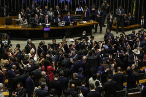 A imagem mostra uma sessão no Congresso Nacional do Brasil, com muitos deputados e senadores interagindo entre si. No fundo, é possível ver algumas pessoas sentadas em mesas, enquanto outras estão em pé, algumas tirando selfies. O ambiente é formal, com muitos dos presentes vestindo ternos escuros. Há bandeiras do Brasil visíveis ao fundo.