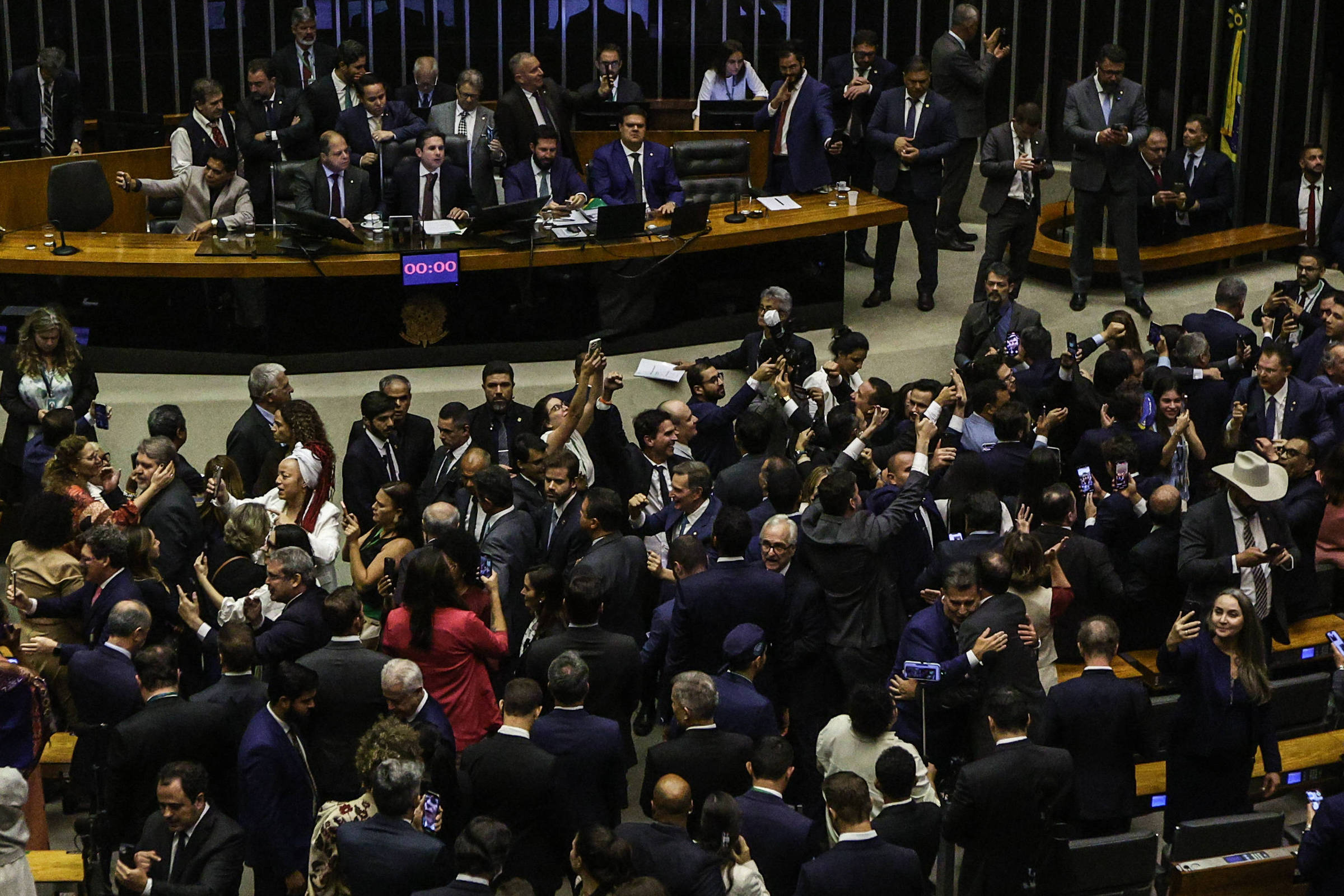 A imagem mostra uma sessão no Congresso Nacional do Brasil, com muitos deputados e senadores interagindo entre si. No fundo, é possível ver algumas pessoas sentadas em mesas, enquanto outras estão em pé, algumas tirando selfies. O ambiente é formal, com muitos dos presentes vestindo ternos escuros. Há bandeiras do Brasil visíveis ao fundo.