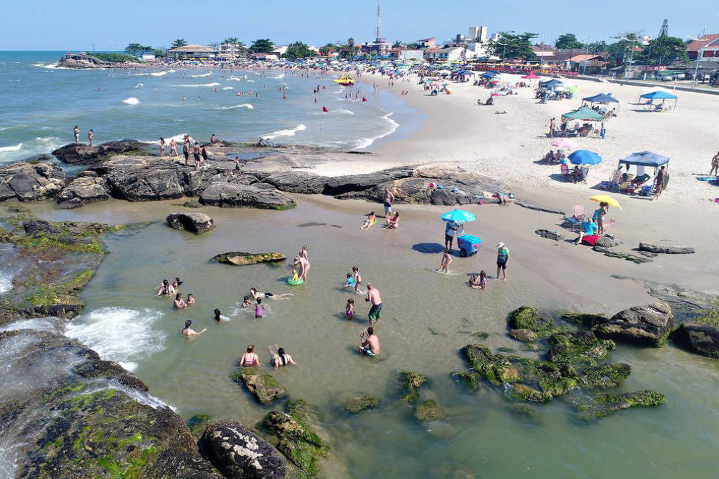 A imagem mostra uma praia com várias pessoas se divertindo. Há um grupo de pessoas na água, algumas sentadas nas rochas e outras brincando na areia. Ao fundo, é possível ver várias barracas e guarda-sóis coloridos, além de um grande número de pessoas na areia. O céu está claro e o mar apresenta ondas suaves.