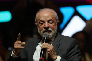 Um homem de cabelos grisalhos e barba, vestido com um terno escuro e uma camisa branca, está falando ao microfone. Ele gesticula com a mão direita, apontando para a audiência, enquanto expressa suas ideias de forma enfática. O fundo é desfocado, mas há luzes coloridas que sugerem um ambiente de evento ou conferência.