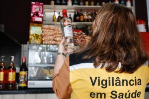Agente da Vigilância em Saúde, vista de costas com colete amarelo e preto, segura uma garrafa de bebida alcoólica em frente a prateleira com diversos produtos e bebidas em um estabelecimento.