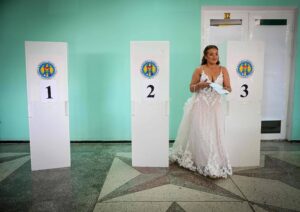 Mulher vestida de noiva segura documentos e está ao lado da cabine de votação número 3 em ambiente interno com paredes verdes.