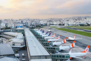 Imagem de um aeroporto com várias aeronaves estacionadas em frente a um terminal. Ao fundo, há uma vista da cidade com prédios altos e um céu com nuvens. A pista de pouso e decolagem é visível, e algumas aeronaves estão em movimento. O ambiente é urbano, com uma mistura de infraestrutura aeroportuária e paisagem urbana