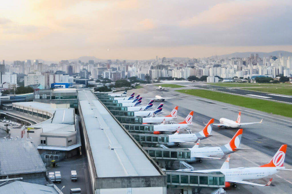 Imagem de um aeroporto com várias aeronaves estacionadas em frente a um terminal. Ao fundo, há uma vista da cidade com prédios altos e um céu com nuvens. A pista de pouso e decolagem é visível, e algumas aeronaves estão em movimento. O ambiente é urbano, com uma mistura de infraestrutura aeroportuária e paisagem urbana