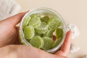 Uma mão segurando um frasco de vidro transparente cheio de doces em forma de folhas, de cor verde. O fundo é de uma tonalidade clara, e há algumas pedras brancas ao redor do frasco. Um pano de tecido claro está parcialmente visível ao fundo.