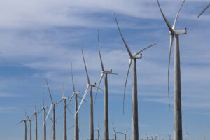 A imagem mostra uma fileira de turbinas eólicas em um campo aberto, com um céu azul e algumas nuvens ao fundo. As turbinas estão alinhadas em uma linha reta, com suas lâminas girando ao vento.