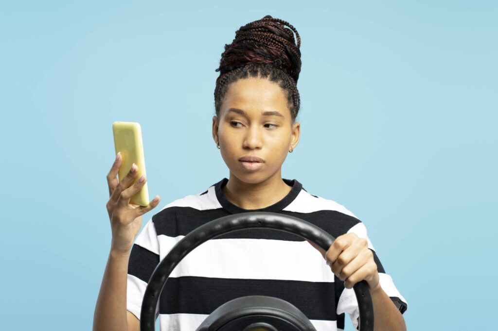 Studio portrait of a young woman gripping a steering wheel while engaging with smartphone