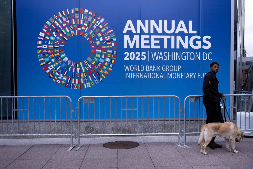 Homem de segurança com cão guia caminha em frente a painel azul com logotipo circular de bandeiras e texto sobre reuniões anuais 2025 do Banco Mundial e FMI em Washington DC.