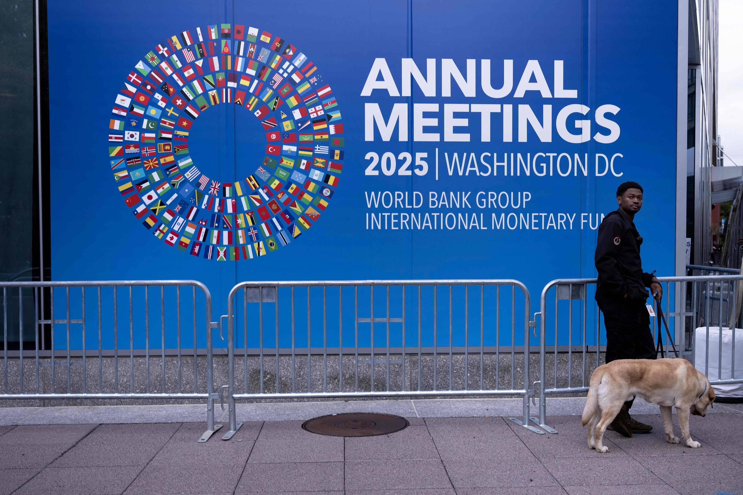 Homem de segurança com cão guia caminha em frente a painel azul com logotipo circular de bandeiras e texto sobre reuniões anuais 2025 do Banco Mundial e FMI em Washington DC.