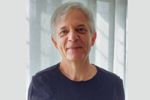 Homem de meia-idade com cabelo grisalho curto, usando camiseta preta, posando em frente a uma cortina branca translúcida com luz natural suave.