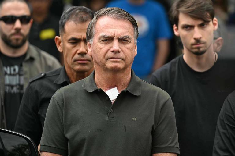 Homem de meia-idade com camisa polo escura está no centro da imagem, com expressão séria. Atrás dele, quatro homens jovens e adultos também vestem roupas escuras, em ambiente externo.