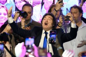 Homem de terno preto e gravata azul levanta os braços e fala animado diante de microfone, cercado por pessoas que filmam e fotografam com celulares e câmeras.