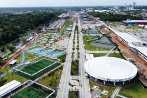 A imagem mostra uma vista aérea de uma área recreativa com várias instalações. Há campos de futebol, piscinas e áreas verdes. No centro, destaca-se uma estrutura circular, possivelmente um centro de eventos. Ao fundo, vê-se uma cidade com prédios e uma área em construção. O céu está claro e a vegetação é densa ao redor.