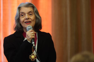 Mulher idosa com cabelos grisalhos e roupas escuras segura microfone e fala. Ela usa uma medalha dourada pendurada no pescoço. Fundo com cortinas em tons de laranja e marrom.