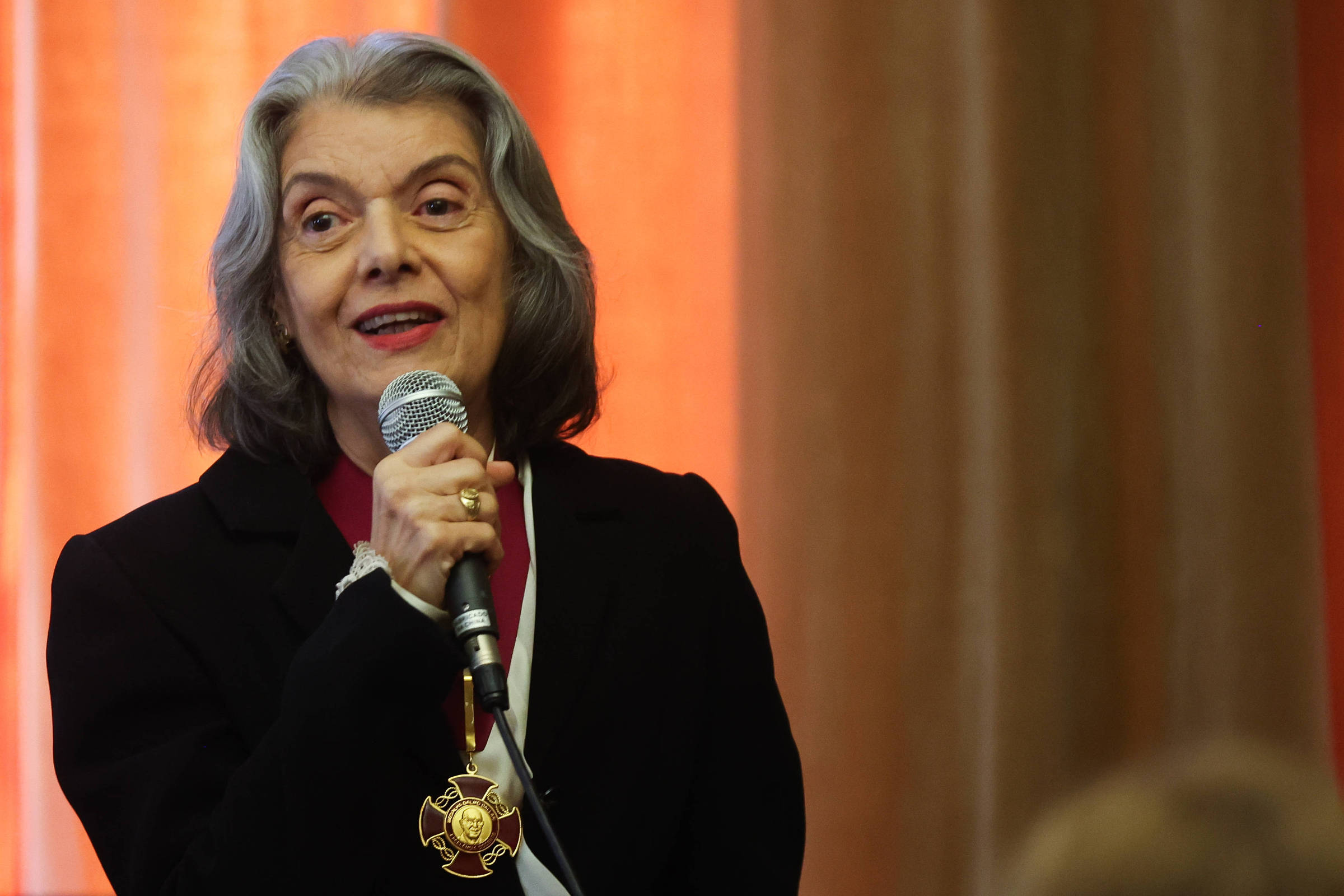 Mulher idosa com cabelos grisalhos e roupas escuras segura microfone e fala. Ela usa uma medalha dourada pendurada no pescoço. Fundo com cortinas em tons de laranja e marrom.
