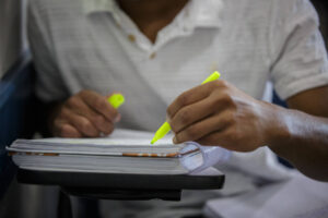 A imagem mostra um estudante em um ambiente de estudo, utilizando uma caneta marca texto amarela para destacar informações em um caderno ou livro. O estudante está com uma camisa branca e as mãos estão posicionadas sobre o material de estudo, que está apoiado em uma superfície.