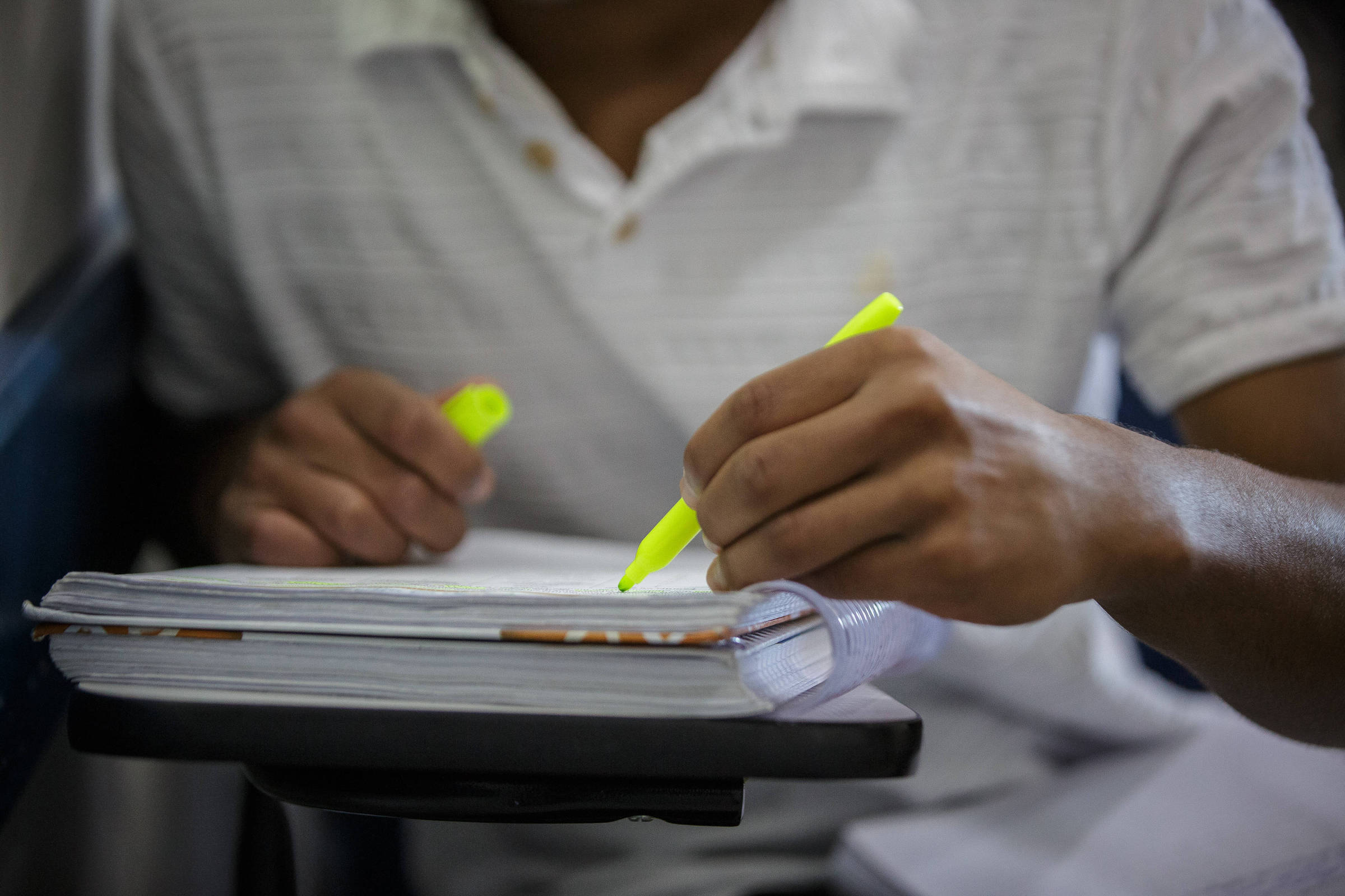 A imagem mostra um estudante em um ambiente de estudo, utilizando uma caneta marca texto amarela para destacar informações em um caderno ou livro. O estudante está com uma camisa branca e as mãos estão posicionadas sobre o material de estudo, que está apoiado em uma superfície.