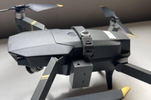 Drone cinza com quatro hélices dobradas, cada uma com ponta amarela, posicionado sobre superfície branca. Estrutura compacta com câmera frontal visível e braços articulados.