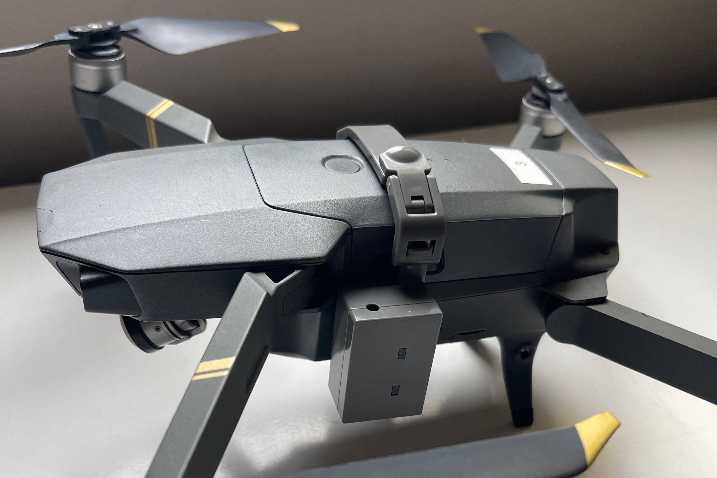 Drone cinza com quatro hélices dobradas, cada uma com ponta amarela, posicionado sobre superfície branca. Estrutura compacta com câmera frontal visível e braços articulados.
