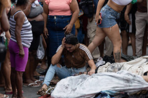 Mulher sentada no chão com expressão de dor ou desespero, rodeada por várias pessoas em pé, algumas observando. No chão, cobertores ou tecidos espalhados, sugerindo situação de emergência ou crise.