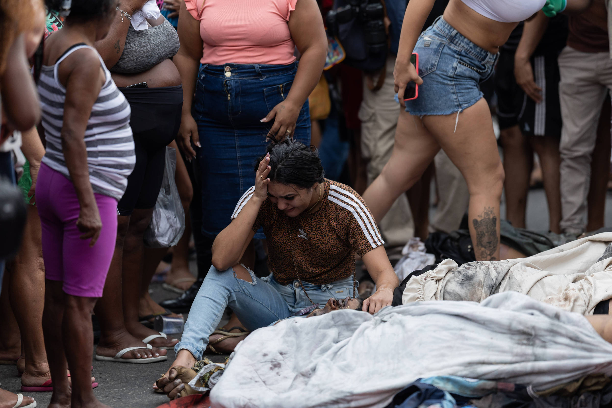 Mulher sentada no chão com expressão de dor ou desespero, rodeada por várias pessoas em pé, algumas observando. No chão, cobertores ou tecidos espalhados, sugerindo situação de emergência ou crise.