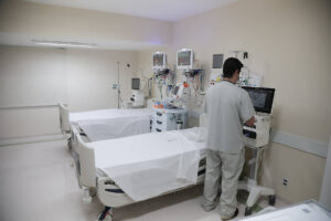 Quarto hospitalar com duas camas de hospital alinhadas, cobertas por lençóis brancos. Profissional de saúde em uniforme branco está de costas, operando um equipamento médico ao lado da cama mais próxima. Monitores e dispositivos médicos estão instalados na parede acima das camas. Ambiente limpo e iluminado, com paredes claras e piso branco.