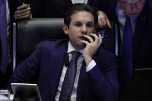 Senador sentado em cadeira preta, vestindo terno azul escuro e gravata roxa, falando ao telefone celular. Dois homens ao redor observam o aparelho, um deles com terno e gravata azul.