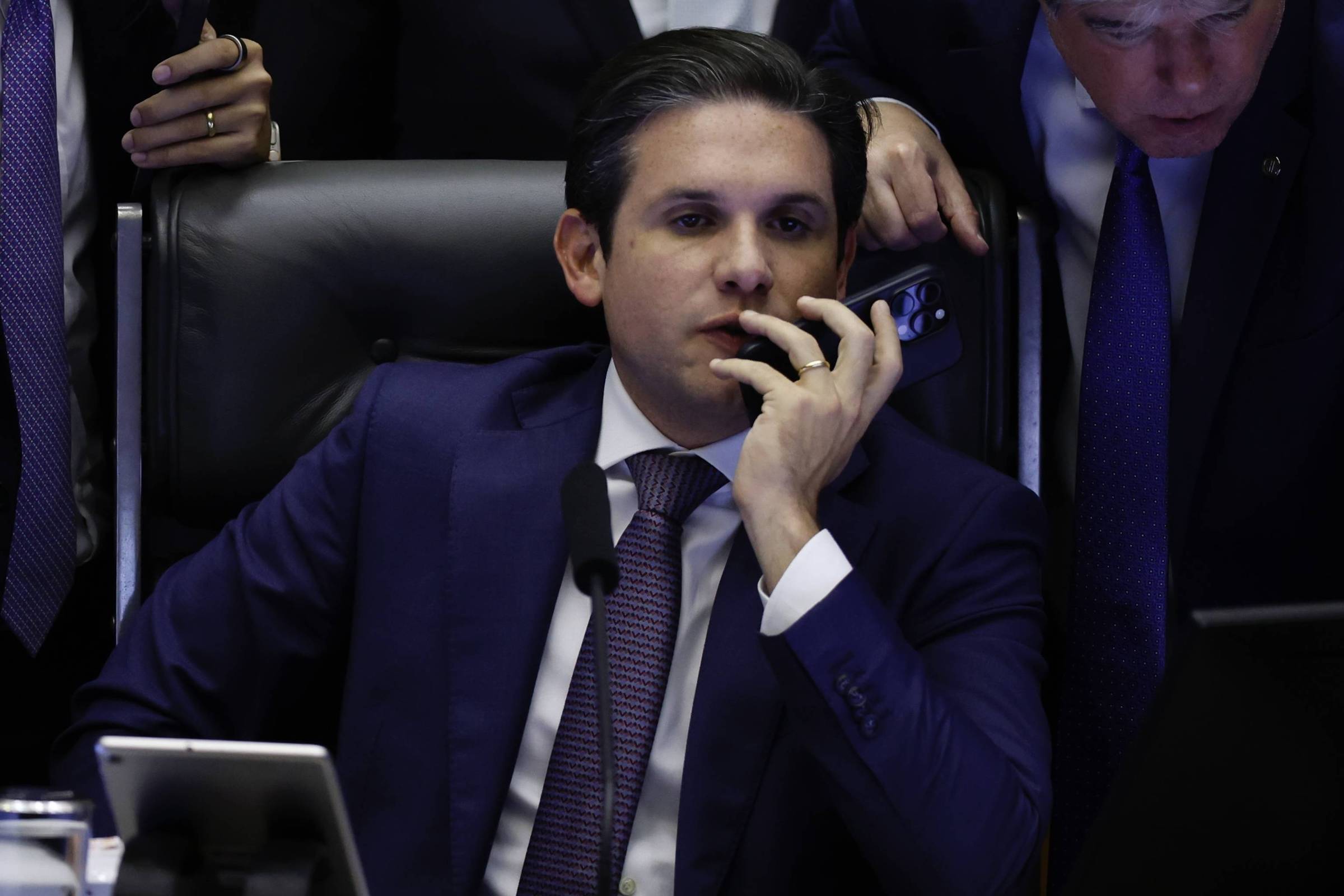 Senador sentado em cadeira preta, vestindo terno azul escuro e gravata roxa, falando ao telefone celular. Dois homens ao redor observam o aparelho, um deles com terno e gravata azul.