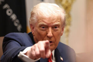 Ex-presidente dos Estados Unidos em terno azul escuro e gravata vermelha, apontando diretamente para a câmera em ambiente interno com bandeira desfocada ao fundo.