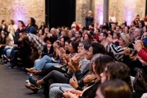 Cerca de 50 pessoas sentadas em cadeiras em ambiente interno com paredes de pedra iluminadas por luzes roxas. O público está voltado para a frente, aplaudindo e prestando atenção ao que ocorre fora do quadro.