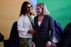 A imagem mostra duas mulheres em um evento. A mulher à esquerda usa óculos, uma blusa verde e um casaco branco, com uma saia estampada. A mulher à direita está vestindo um vestido xadrez e parece sorrir enquanto a outra mulher a toca levemente nas costas. Ao fundo, há uma parede com cores em degradê.